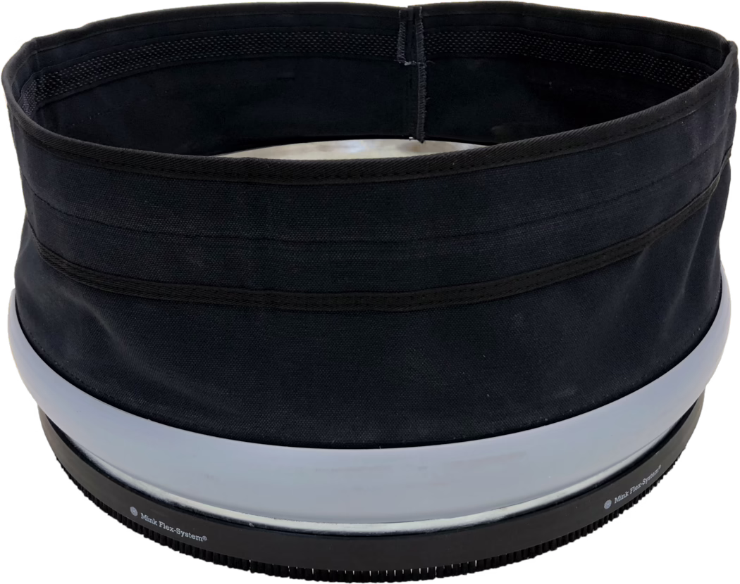 Close-up view of the top rim and opening of a cylindrical black fabric container or hat. The image shows a circular inner opening with black lining, visible reinforced stitching and several horizontal fabric bands on the exterior. Near the lower edge there are two wide light-gray plastic rings and a black ribbed rubber base with small teeth and partially visible printed text 'Mink Flex-System®'. The fabric has a coarse weave and a matte finish; the object appears structured yet flexible. Lighting is even and the background is white.